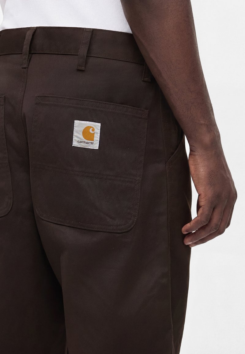 Pantalon Carhartt marron foncé avec une grande poche arrière ornée d'un patch logo Carhartt blanc et orange, porté par une personne vêtue d'un haut blanc.
