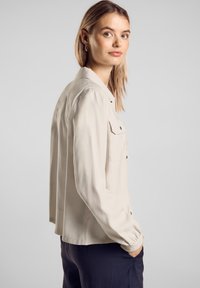 Vaaleanbeige nappipaita, jossa on kaksi etutaskua, pitkät hihat ja muotoiltu malli. Sileä rakenne ja hillitty kiilto.