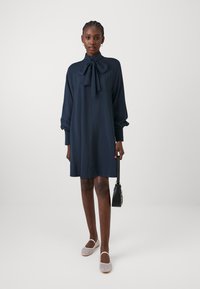 Sininen mekko, jossa on solmion kaulus, pitkät hihat ja istuvat hihansuut; yhdistetty vaaleankeltaisiin kudottuihin kenkiin ja pieneen mustaan käsilaukkuun.