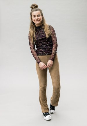 Langarm, aangesloten bovenkleding met een paars en gouden patroon; beige flarebroeken; zwart-witte sneakers; model glimlachend.