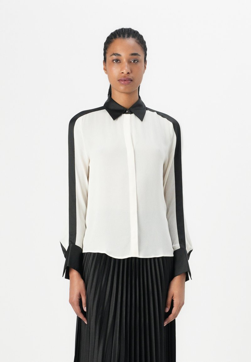 Blouse blanche avec des accents noirs, dotée d'un col, d'une fermeture à boutons sur le devant et de manches longues. Texture lisse, couleur unie et design structuré.