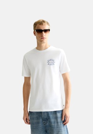 Young man wearing sunglasses, a white t-shirt with small text on the chest, and blue denim jeans against a plain white background.