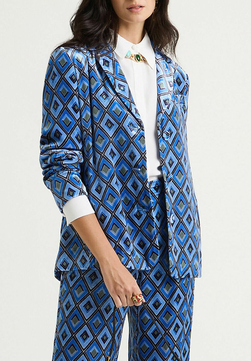 Costume à motifs bleus avec un design en losange dans différentes nuances de bleu et vert. Le blazer a une coupe cintrée avec un revers cranté.