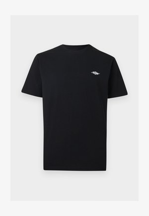 Giovane uomo con capelli corti e scuri che indossa una t-shirt nera con un piccolo logo bianco sul petto e jeans scuri, rivolto in avanti su sfondo bianco.