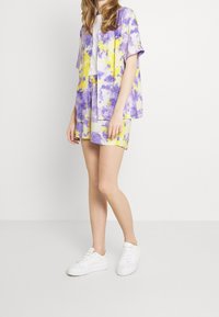 Completo composto da camicia a maniche corte e pantaloni corti in un design floreale viola e giallo, realizzato in tessuto leggero, abbinato a sneaker bianche.