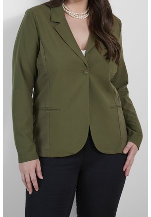 Vrouw draagt een olijfgroene blazer met één knoop, zwarte broek, wit topje en parelketting, staande tegen een witte achtergrond.