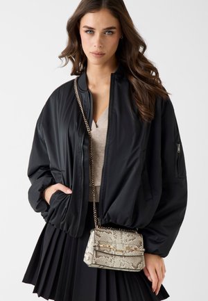 Frau trägt eine schwarze Bomberjacke, ein beigefarbenes Oberteil, einen schwarzen Faltenrock und eine Umhängetasche mit Schlangenledermuster und goldener Kettenriemen.