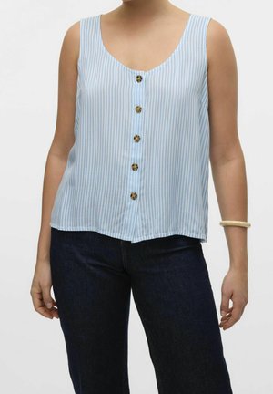 Kobieta w bezrękawowej bluzce w jasnoniebieskie i białe pionowe paski z pięcioma guziczkami z przodu, ciemnych dżinsach i kremowej bransoletce na nadgarstku.