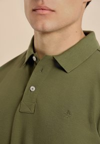 Polo en verde oliva hecho de tela texturizada, con cuello, tres botones y un logo bordado en el pecho.