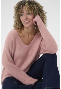 Pulover tricotat roz deschis, cu decolteu în V, textură cu rimă și mâneci lungi. Modelul poartă blugi albastru închis, așezat într-o poziție relaxată.