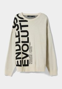 Pull en maille beige clair avec des graphismes en texte noir. Coupe oversize avec un col rond, des poignets et un ourlet côtelés. Texture douce.