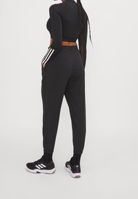Zwart sportoutfit met een cropped lange mouw top en aansluitende broek. De broek heeft zijzakken en witte strepen. Sneakers zijn zwart.