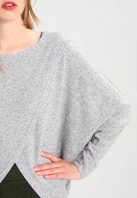 Szary, luźno dopasowany sweter wykonany z miękkiej dzianiny, z szerokim dekoltem oraz rękawami typu dolman, z wyraźnym dnem i ribbowymi detalami.