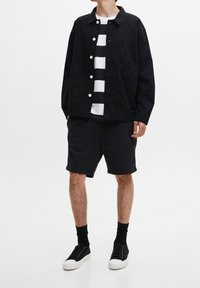 Schwarze Hemdjacke über einem weiß-schwarz gestreiften Shirt, kombiniert mit schwarzen Shorts und schwarzen Socken, abgerundet mit schwarzen Sneakers.