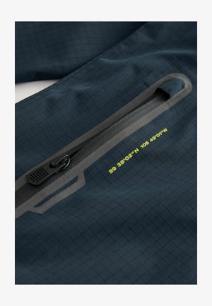 Tissu bleu foncé présentant une texture en grille, avec une poche zippée noire et des accents réfléchissants. Des coordonnées imprimées jaunes sont visibles.