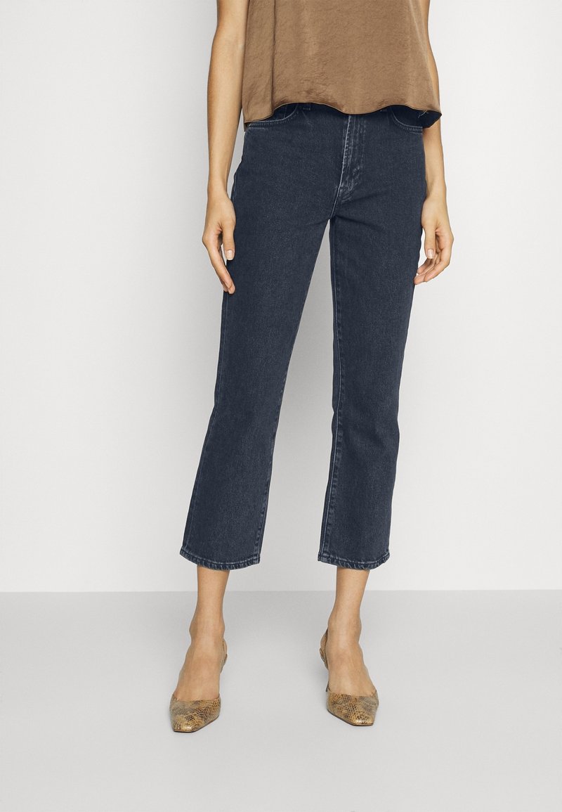 Femme portant un haut sans manches marron, un jean bleu foncé raccourci et des ballerines marron à bout pointu, debout devant un fond uni.