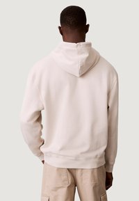 Felpa con cappuccio beige in morbido cotone, caratterizzata da un grande cappuccio posteriore, polsini e orlo a costine. Il tessuto ha una texture liscia con cuciture minime.