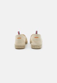 Chaussures slip-on en toile beige avec une semelle en espadrille en jute tissé. L'accent à l'arrière comprend une boucle rayée rouge, blanche et bleue.