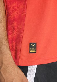 Maillot de football rouge avec un tissu texturé, présentant un panneau latéral à motifs et une étiquette de marque noire indiquant "LICENCE DE FOOTBALL AUTHENTIQUE."