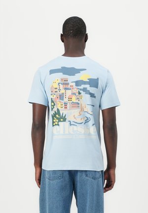 Mężczyzna w jasnoniebieskiej koszulce z kolorową grafiką nadmorskiego miasteczka i logo ellesse na plecach, w połączeniu z niebieskimi dżinsami.
