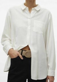 Valkoinen kauluksellinen nappipaita, jossa on rintatasku, pitkät hihat ja hieman väljä istuvuus, yhdistetty mustiin housuihin ja beigeen vyöhön.