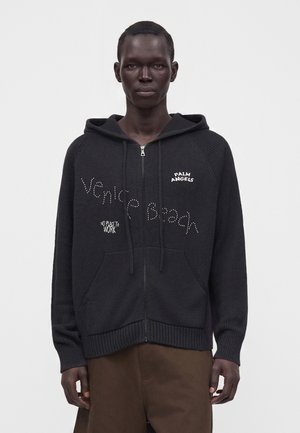 Mężczyzna w czarnej bluzie z kapturem na zamek, z tekstem "Venice Beach," "Palm Angels," oraz "No place to work" na przodzie.