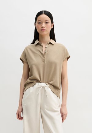 Fiatal nő, bézs rövid ujjú galléros pólót és laza, fehér zsinóros nadrágot visel, egyszínű fehér háttér előtt áll.