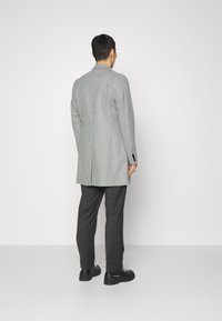Šedý blazer ze směsi vlny s hladkou texturou, s klopami, dvěma zadními ventilačními otvory a černými knoflíky, doplněný tmavými kalhotami a černou obuví.