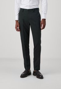 Dark green tailored pants with a smooth texture, featuring a button closure and side pockets, paired with black leather shoes.