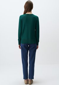 Zielony sweter z długim rękawem ma okrągły dekolt, dopasowane mankiety i nieco dłuższy tylny brzeg, zestawiony z niebieskimi spodniami typu straight-leg.