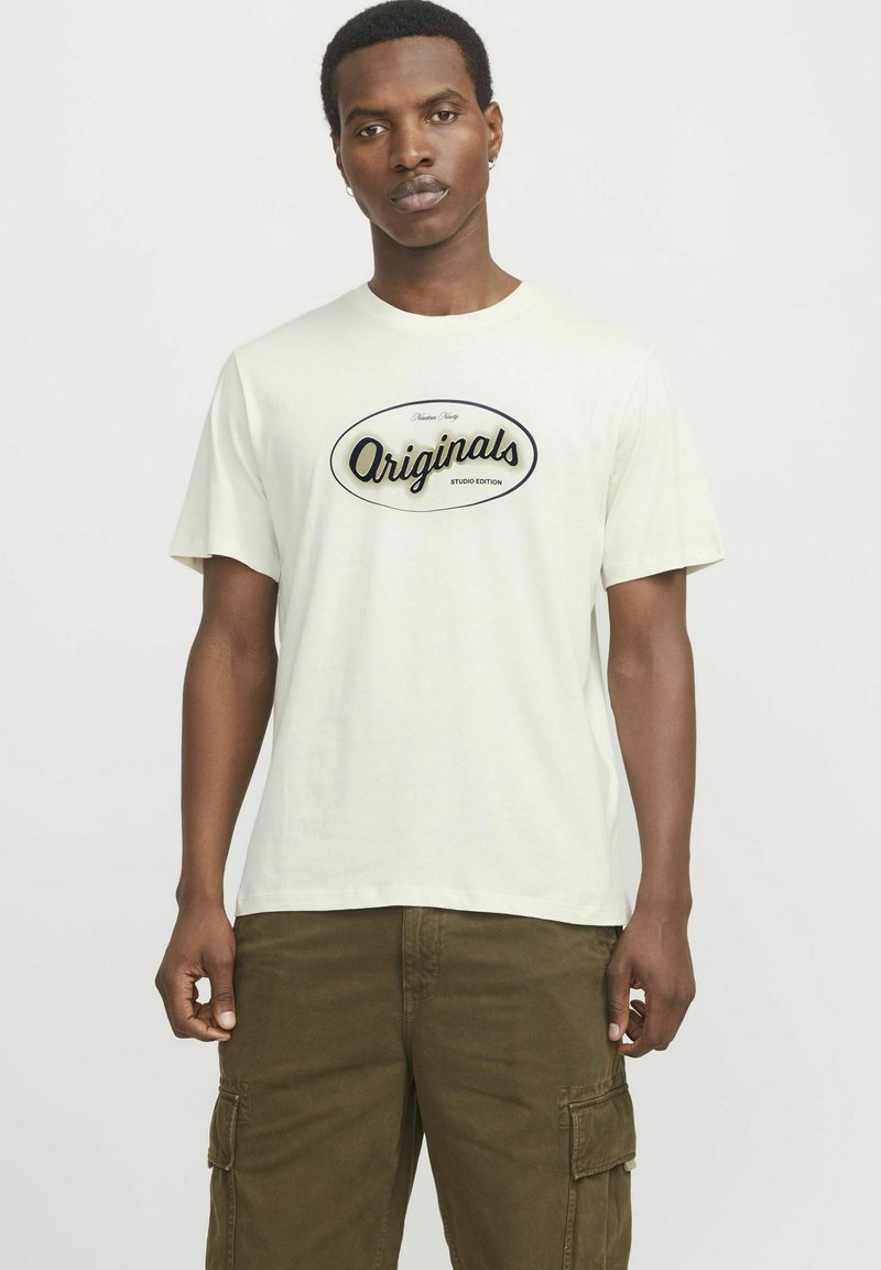 Hellbeige Baumwoll-T-Shirt mit kurzen Ärmeln und Rundhalsausschnitt, das auf der Vorderseite ein schwarzes "Originals"-Logo in ovaler Umrandung zeigt.