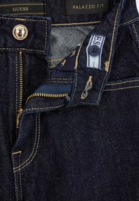 Jeans in denim blu scuro con zip dorata, etichette di marca e cuciture a contrasto. Presentano una vestibilità palazzo e chiusura con bottone.
