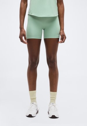 Person wearing light green athletic shorts, beige crew socks, and white sneakers standing against a plain white background.