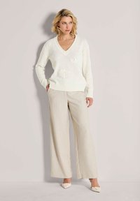 Vrouw in een witte V-hals trui met bloemmotieven, beige wijde broek en witte puntige hakken, staand tegen een lichte achtergrond.