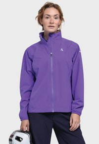 Frau, die eine lila Zip-Jacke und dunkle Hosen trägt, hält mit ihrer linken Hand einen weißen Helm mit schwarzen Details.