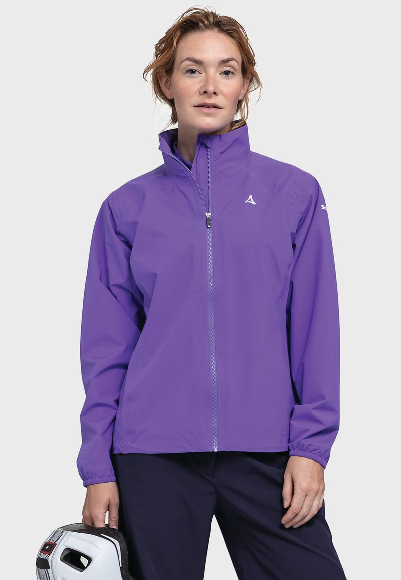 Frau, die eine lila Zip-Jacke und dunkle Hosen trägt, hält mit ihrer linken Hand einen weißen Helm mit schwarzen Details.