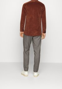 Hnědá kordurová košile s hladkým povrchem, dlouhými rukávy a knoflíkovými manžetami, spárována se světle šedými dobře padnoucími kalhotami a bílými teniskami.