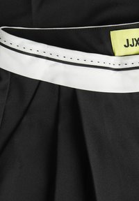 Czarna tkanina z białym paskiem i wykończeniem, z fałdami i metką z napisem "JJX" w kolorze żółtym. Materiał wydaje się gładki i strukturalny.