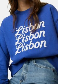 Mujer con un sudadera azul que tiene "Lisboa" en una escritura blanca repetida tres veces y unos jeans de tiro alto en azul claro.
