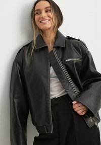 Giacca bomber in pelle nera con colletto a pressione, tasche con zip e polsini a costine. Presenta una vestibilità rilassata e una texture liscia.