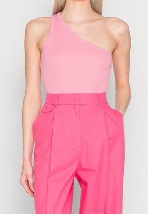 Vrouw draagt een mouwloze roze een-schouder top en hoog getailleerde felroze op maat gemaakte broek met handen in de zakken.