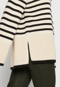 Sweter w paski w kremowym i czarnym kolorze, z prążkowaną teksturą, bocznymi rozcięciami oraz luźnym krojem, z kontrastowymi ściągaczami i dołem.