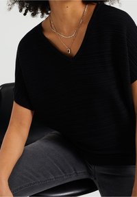 Černý pletený top s ribkovaným vzorem, V- výstřihem a krátkými rukávy, v kombinaci se šedými džíny. Přidané zlaté náhrdelníky v několika vrstv ách dodávají styl.