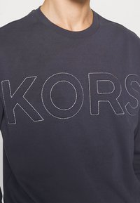 Tmavě modrý sweatshirt s kulatým výstřihem, na přední straně s oversized bílým vyšitým nápisem "KORS". Měkká textilie.