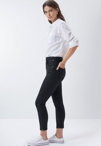 Jeans skinnys negros de denim, con cintura alta, bolsillos frontales y cierre de cremallera. Combinados con una camisa blanca de botones y zapatillas blancas.