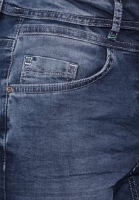 Jeans de mezclilla azul oscuro con una textura suave, costuras detalladas y un pequeño bolsillo delantero con costuras de acento en verde en contraste.