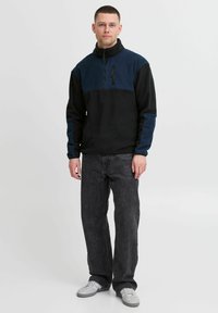 Black and navy fleece pullover with a zip neck, long sleeves, and front pockets. Worn with dark gray jeans and white sneakers.