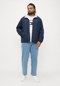 Jachetă de vânt bleumarin cu guler gri, asortată cu un tricou alb cu imprimeu și blugi albastru deschis. Modelul poartă adidași albi.