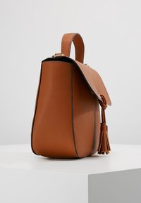 Hnědá kožená kabelka s horním uchopem, klopovým zavíráním a ozdobnou třásní, zobrazená z bočního úhlu na bílé ploše.