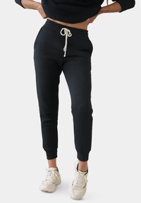 Zwarte sweatpants met elastische boorden en tailleband, voorzien van een crèmekleurige koord en een gladde textuur, gecombineerd met lichtgekleurde sneakers.